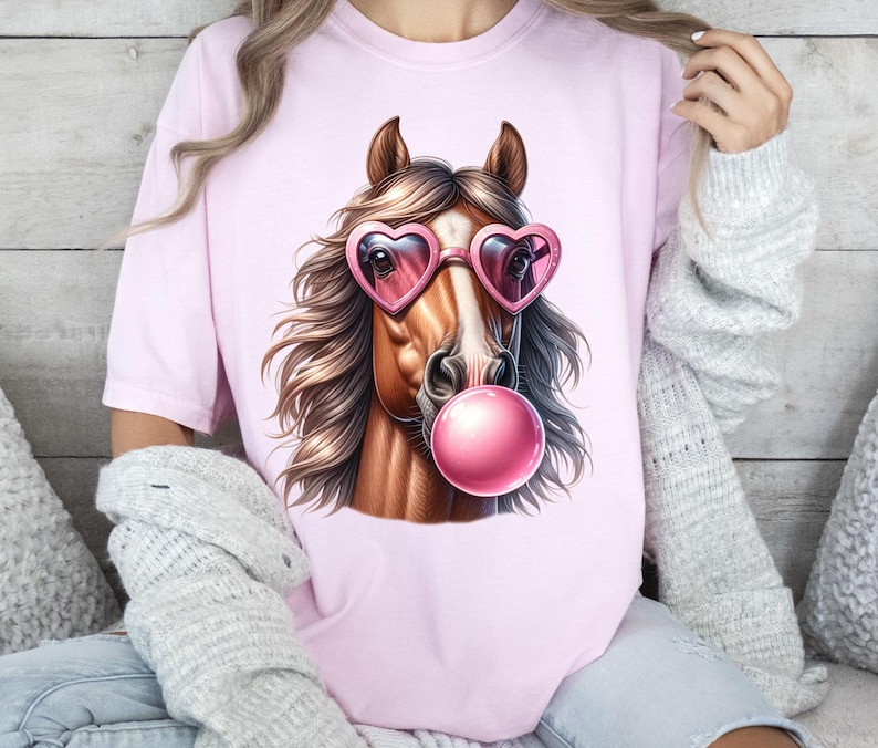 May include: A pink t-shirt featuring a graphic of a brown horse wearing pink heart-shaped sunglasses and blowing a pink bubble.