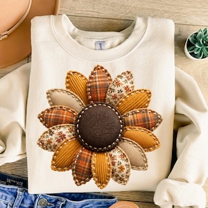 Könnte beinhalten: Ein cremefarbenes Sweatshirt mit Rundhalsausschnitt und einer großen Sonnenblumen-Grafik in der Mitte. Die Blütenblätter bestehen aus verschiedenen Stofftexturen wie Cord, Karomustern und Blumenprints, die mit sichtbaren Ziernähten versehen sind.