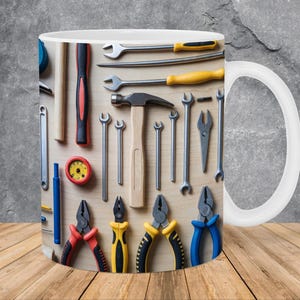 May include: White ceramic mug featuring a detailed print of various hand tools, including wrenches, pliers, a hammer, and screwdrivers, arranged on a wooden surface. The tools are in shades of silver, black, red, and yellow.