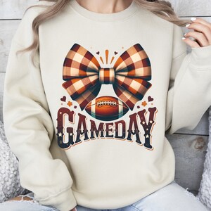 May include: A cream-colored sweatshirt with a brown and white plaid bow, a football, and the text "GAMEDAY" in a vintage style font.