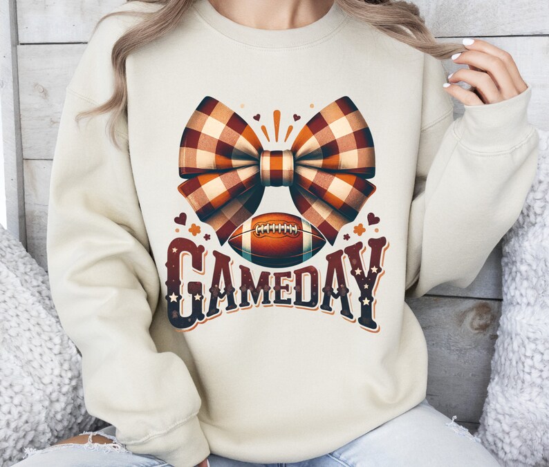 May include: A cream-colored sweatshirt with a brown and white plaid bow, a football, and the text "GAMEDAY" in a vintage style font.