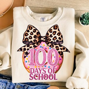 Puede incluir: Sudadera color crema con un diseño de donut rosa con chispas y el texto "100 Days of School" en purpurina rosa. Un gran lazo con estampado de leopardo está encima del donut. La sudadera es de estilo casual.