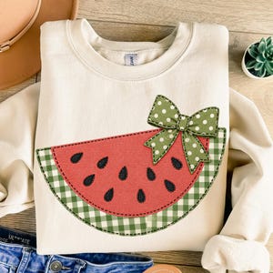 May include: A cream-colored crewneck sweatshirt featuring a large applique of a watermelon slice. The watermelon has a red center with black seeds, a green and white gingham rind, and a green bow with white polka dots attached to the top.