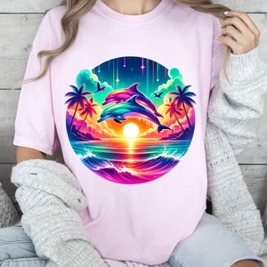 May include: A pink t-shirt with a colorful graphic of two dolphins jumping over a sunset with palm trees and a rainbow sky.