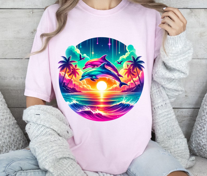 May include: A pink t-shirt with a colorful graphic of two dolphins jumping over a sunset with palm trees and a rainbow sky.