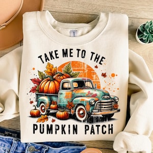 May include: Cream-colored sweatshirt featuring a fall-themed graphic. The design includes a vintage teal truck filled with pumpkins, surrounded by autumn leaves, and the text "TAKE ME TO THE PUMPKIN PATCH."
