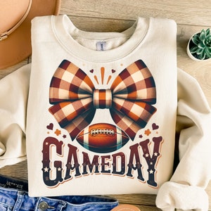 May include: A cream-colored sweatshirt with a brown and white plaid bow, a football, and the text "GAMEDAY" in a vintage style font.