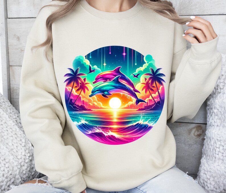 May include: A cream-colored sweatshirt with a colorful graphic of two dolphins jumping over a sunset ocean scene. The graphic includes palm trees, stars, and a rainbow-colored wave.