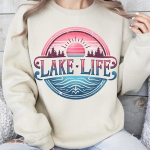 May include: A cream-coloured sweatshirt with a vintage-style graphic design featuring a sunset, trees, and the text "LAKE LIFE" in a blue, pink, and white colour scheme.
