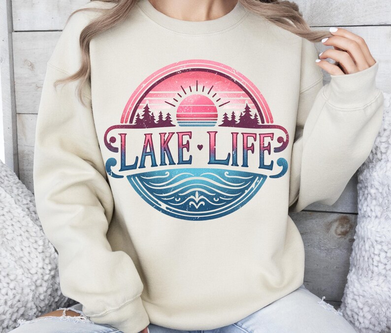 May include: A cream-coloured sweatshirt with a vintage-style graphic design featuring a sunset, trees, and the text "LAKE LIFE" in a blue, pink, and white colour scheme.