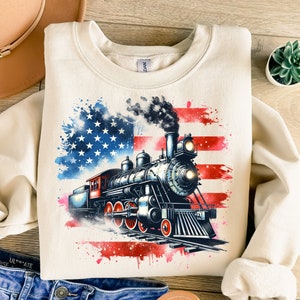 May include: A cream-colored sweatshirt with a watercolor-style American flag design featuring a vintage steam locomotive. The locomotive is black with red accents and is surrounded by red, white, and blue paint splatters.