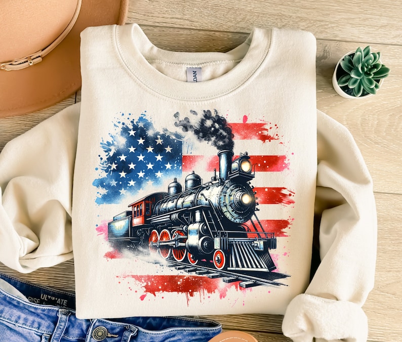 May include: A cream-colored sweatshirt with a watercolor-style American flag design featuring a vintage steam locomotive. The locomotive is black with red accents and is surrounded by red, white, and blue paint splatters.