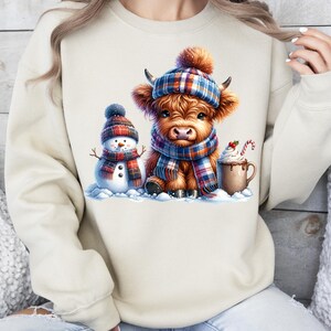 May include: A beige sweatshirt featuring a winter scene with a Highland cow wearing a plaid hat and scarf, a snowman, and a mug of hot chocolate. The cow and snowman are in a snowy setting, with a candy cane in the mug.