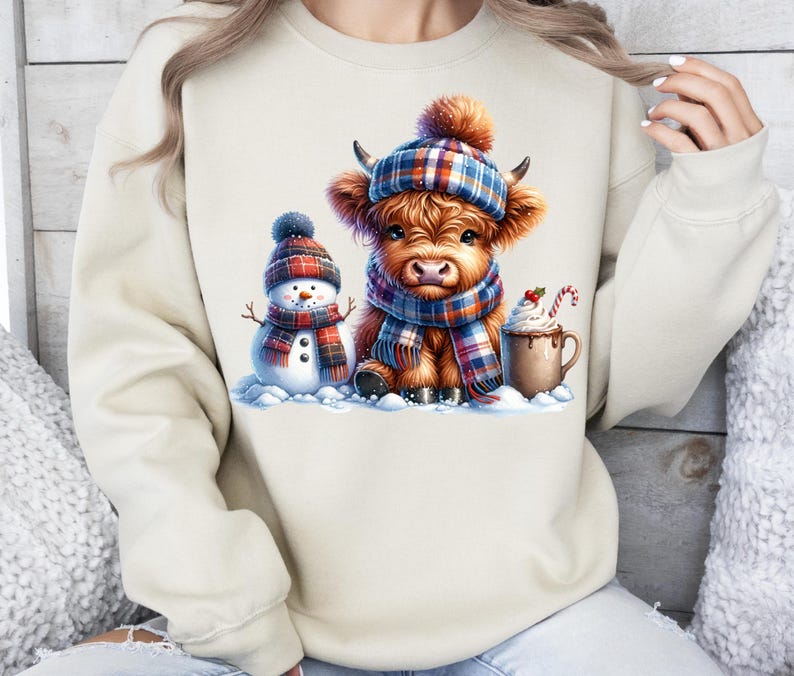 May include: A beige sweatshirt featuring a winter scene with a Highland cow wearing a plaid hat and scarf, a snowman, and a mug of hot chocolate. The cow and snowman are in a snowy setting, with a candy cane in the mug.