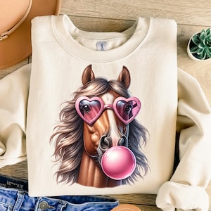 May include: A cream-coloured sweatshirt featuring a graphic of a brown horse wearing pink heart-shaped sunglasses and blowing a pink bubble.