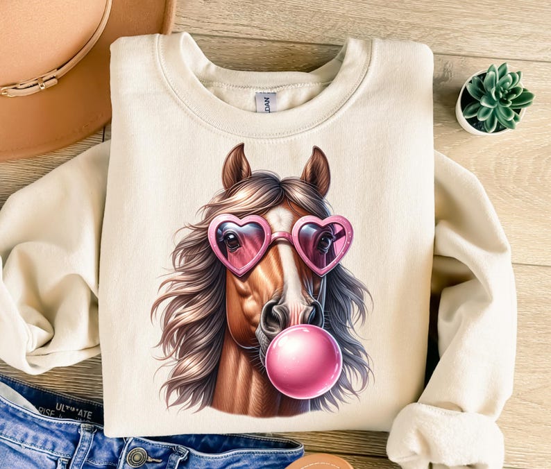 May include: A cream-coloured sweatshirt featuring a graphic of a brown horse wearing pink heart-shaped sunglasses and blowing a pink bubble.