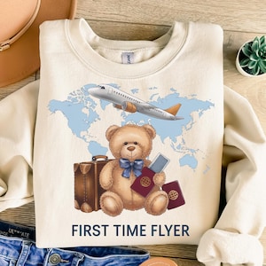 Könnte beinhalten: Ein cremefarbener Pullover mit einem Aufdruck eines Teddybären, der einen Pass und Gepäck hält, ein Flugzeug, das über eine Weltkarte fliegt, und dem Text "FIRST TIME FLYER".