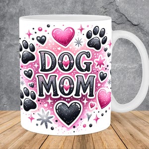 Op de afbeelding: Witte keramische mok met een roze en zwart ontwerp. De mok heeft de tekst "DOG MOM" omringd door harten, sterren en poot afdrukken.