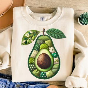 Puede incluir: Sudadera de color crema con un diseño detallado de aguacate. El aguacate está abierto, revelando un hueso marrón y un mosaico de telas verdes. El diseño incluye hojas y un tallo, creando un gráfico vibrante con temática de alimentos.