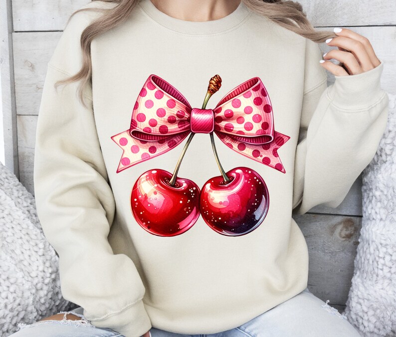 May include: A cream-colored sweatshirt with a pink and white polka dot bow on top of two red cherries.