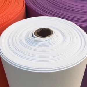 May include: Close-up of a roll of white material with a cardboard core. The material is tightly wound, showing concentric circles. Rolls of orange and purple material are visible in the background.
