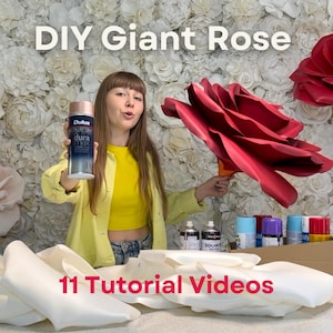 May include: A woman holds a can of spray paint and a large red paper rose. The image has the text "DIY Giant Rose" and "11 Tutorial Videos". Several cans of spray paint and white foam rose petals are visible.