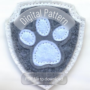 Op de afbeelding: Een grijs en lichtblauw vilten pootafdrukpatch met de woorden "Digital Pattern" en "PDF file to download". De patch is schildvormig met een lichtblauwe pootafdruk in het midden.