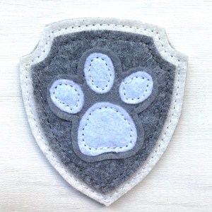 May include: A grey felt patch in the shape of a shield with a white felt paw print in the centre. The paw print has a white stitched outline.