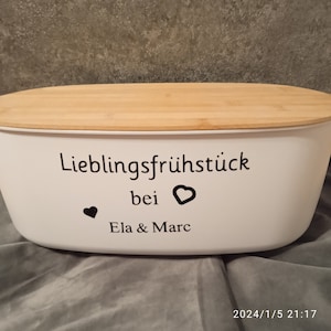 Könnte beinhalten: Ein weißer Brotbehälter aus Keramik mit einem Holzdeckel. Der Behälter hat eine schwarze Aufschrift mit der Aufschrift "Lieblingsfrühstück bei Ela & Marc" mit zwei schwarzen Herzen.