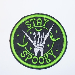 May include: A black and green embroidered patch featuring a white skeleton hand making the 'rock on' gesture. The patch is surrounded by the text 'STAY SPOOKY'.