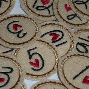 May include: A collection of beige felt circles with brown trim, each featuring a black number with a red heart. The numbers range from 1 to 5.