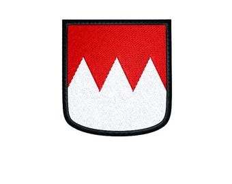 May include: Embroidered patch featuring a shield design with a red upper section and a white lower section with three triangular peaks. The patch has a black border and is likely for clothing or accessories.