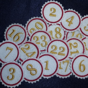 Könnte beinhalten: Ein Satz von vierundzwanzig weißen Filzkreisen mit roten Rändern und goldenen Zahlen von 1 bis 24. Die Kreise sind in einem zufälligen Muster angeordnet.