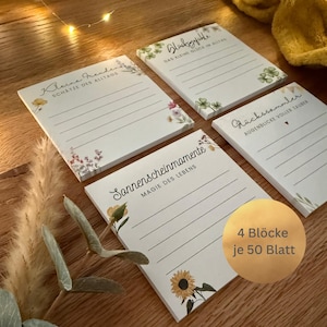 Könnte beinhalten: Vier Notizblöcke mit floralen Designs und linierten Seiten. Jeder Notizblock hat einen anderen deutschen Satz, darunter "Kleine Freuden" und "Sonnenscheinmomente". Die Notizblöcke sind weiß mit floralen Akzenten.