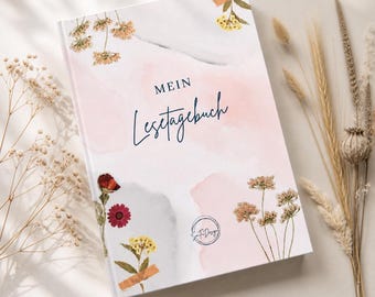 May include: A hardcover journal with the words "Mein Lesetagebuch" in elegant script. The cover features a watercolor design with pink and gray hues, adorned with dried flowers and botanical elements. The journal has a soft, feminine aesthetic.