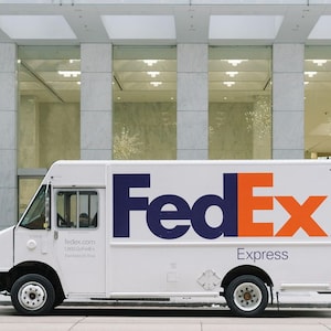 以下が含まれることがあります： 青とオレンジのFedExロゴが書かれた白いFedExの配送トラック。トラックは、大きな窓のある近代的なオフィスビルの前に駐車されています。