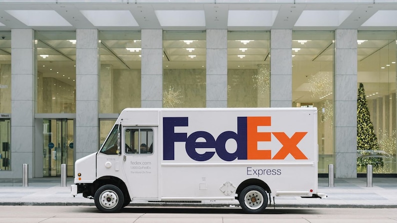 以下が含まれることがあります： 青とオレンジのFedExロゴが書かれた白いFedExの配送トラック。トラックは、大きな窓のある近代的なオフィスビルの前に駐車されています。