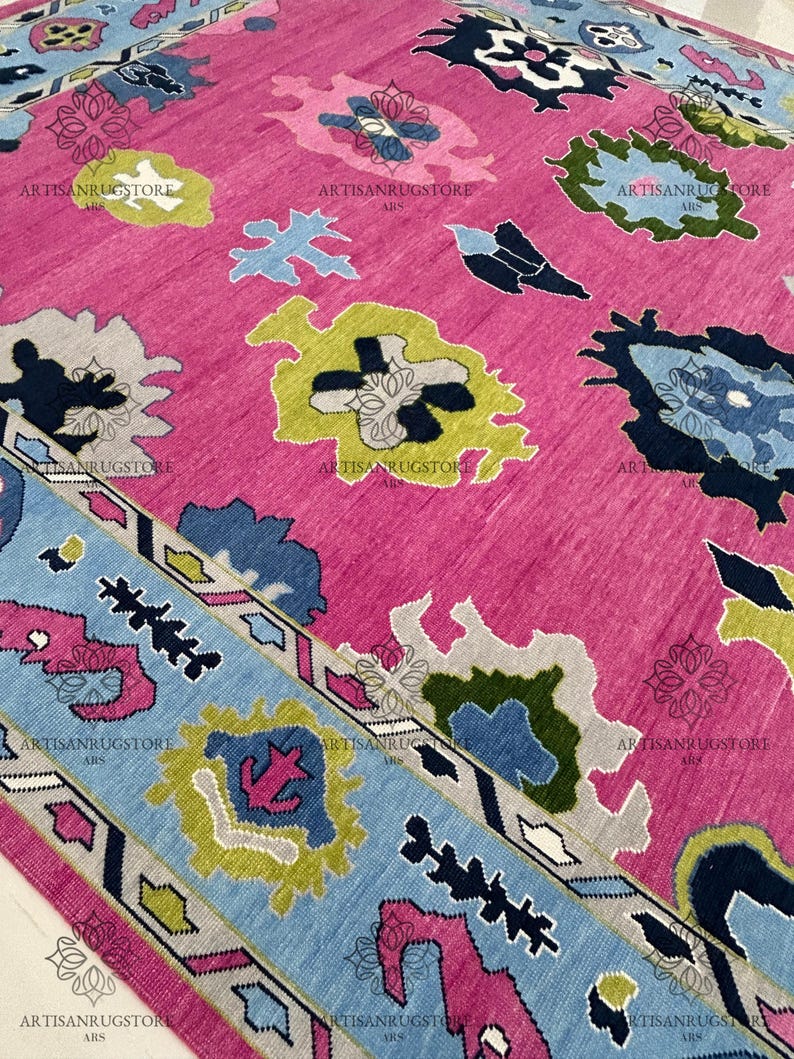 May include: A pink rug with a blue, green, and yellow geometric pattern. The rug has a border with a repeating pattern of triangles and zigzags.