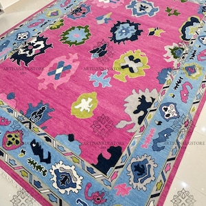 May include: A pink rug with a blue, green, and white geometric pattern. The rug has a border with a repeating pattern of triangles and diamonds.
