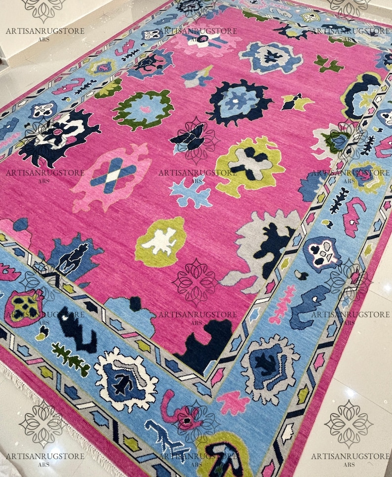 May include: A pink rug with a blue, green, and white geometric pattern. The rug has a border with a repeating pattern of triangles and diamonds.