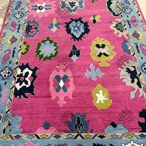 May include: A pink rug with a blue, green, and yellow geometric pattern. The rug has a border with a repeating pattern of triangles and diamonds.