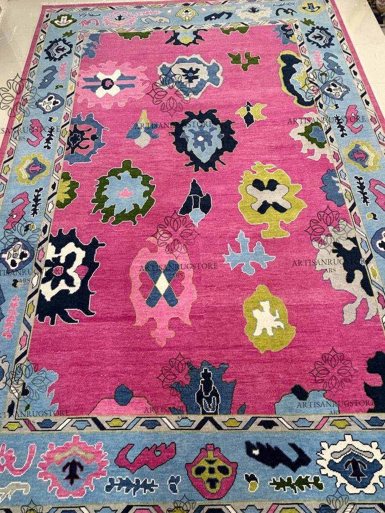 May include: A pink rug with a blue, green, and yellow geometric pattern. The rug has a border with a repeating pattern of triangles and diamonds.