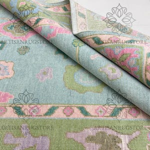 May include: A rolled-up area rug with a floral pattern in shades of blue, pink, green, and purple. The rug features a border with a repeating design and the text "ARTISANRUGSTORE". The rug is made of a woven material.