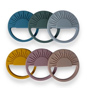 May include: Six silicone teethers shaped like suns with a raised sunburst design. The teethers are in various colors, including blue, green, gray, yellow, pink, and brown.