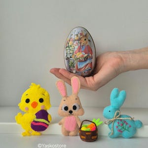 May include: An Easter-themed display featuring felt figurines: a yellow chick holding a decorated egg, a tan bunny, a blue bunny, and a basket of carrots. An egg-shaped tin with a bunny illustration is held in a hand. The text "Vintage Bunny Girl" is visible.