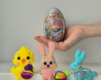 Op de afbeelding: Een Paas-thema display met vilten figuren: een gele kuiken met een versierd ei, een bruin konijn, een blauw konijn en een mandje met wortelen. Een eivormige blik met een konijnenillustratie wordt in een hand gehouden. De tekst "Vintage Bunny Girl" is zichtbaar.
