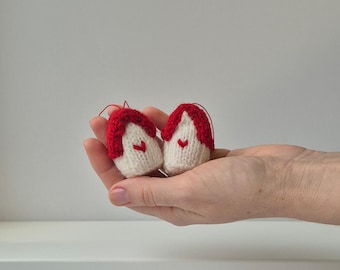Op de afbeelding: Twee kleine, handgebreide huisornamenten. Elk ornament heeft een witte behuizing, een rood dak en een klein rood hart. Ze worden in een hand gehouden tegen een witte achtergrond. Ideaal voor vakantiedecoratie.