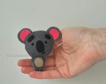 May include: A gray felt koala bear with pink inner ears, black eyes, and a black nose. The koala has a tan belly and is held in a hand. The Etsy shop name "Yaskostore" is in the lower left corner.