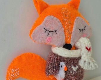 Op de afbeelding: Een handgemaakte oranje vilten vospop met gesloten ogen, gekleed in een bruine trui en een witte sjaal. De vos houdt een kleine witte vogel vast. De pop heeft een witte staart en staat op een wit oppervlak.