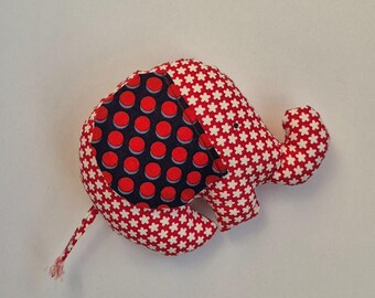 Può includere: Un elefante di stoffa rosso e bianco con una toppa blu e rossa a pois sul dorso. L'elefante ha una piccola coda e un orecchio cadente.